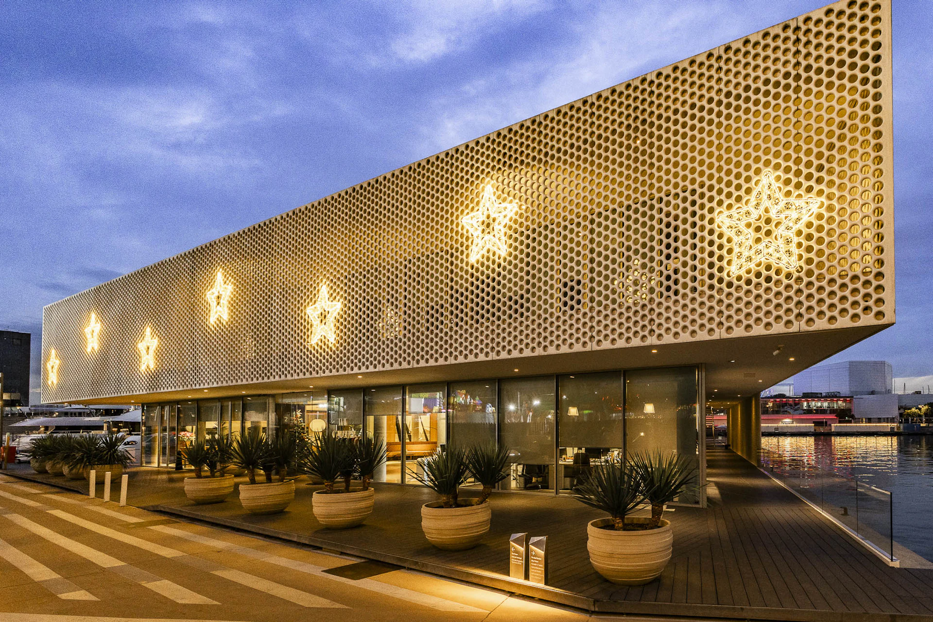 Edificio moderno junto al mar con fachada dorada perforada y patrones de estrellas iluminados al anochecer.