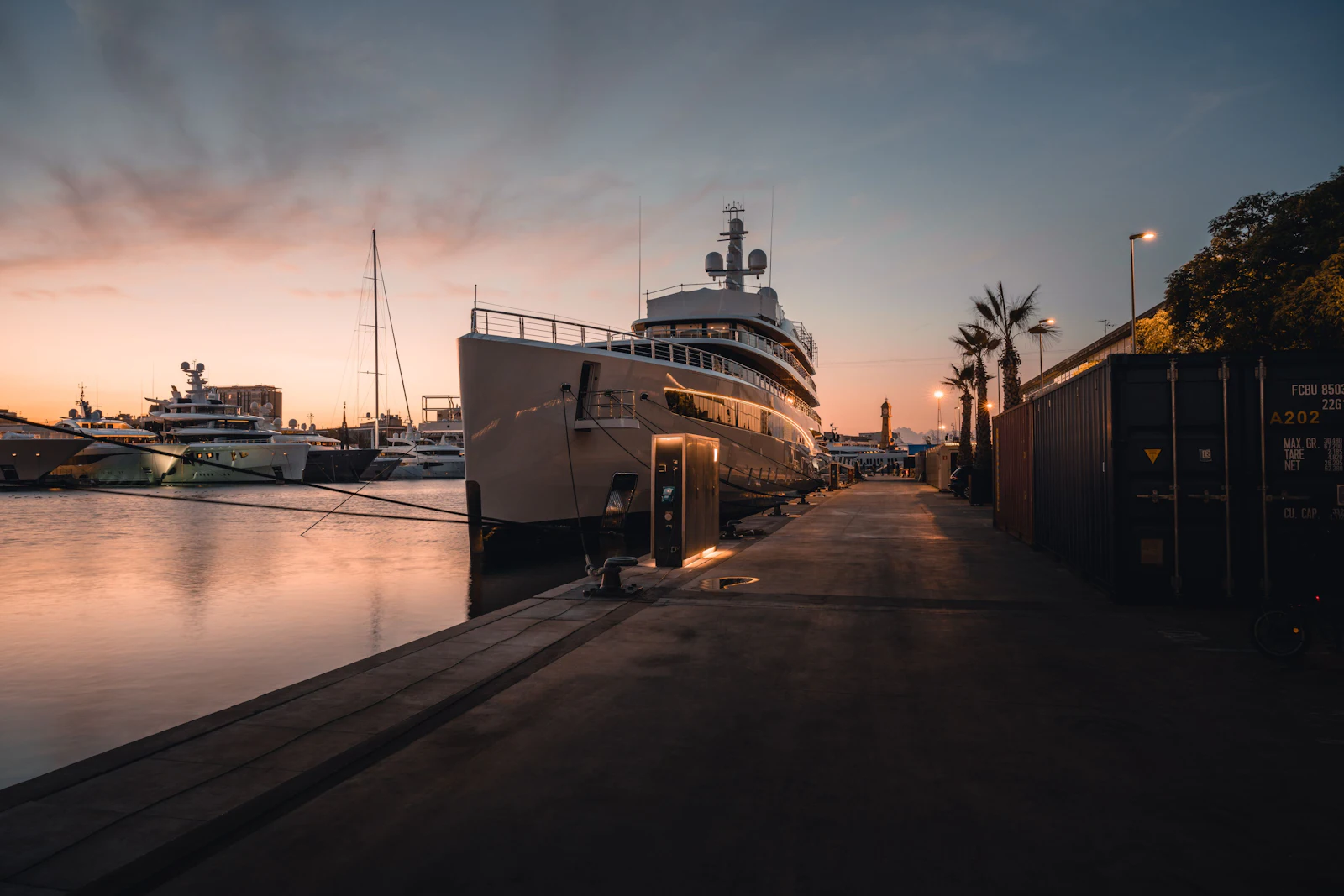 Superyachts moored at a marina pier during sunset with Barcelona cityscape in background.