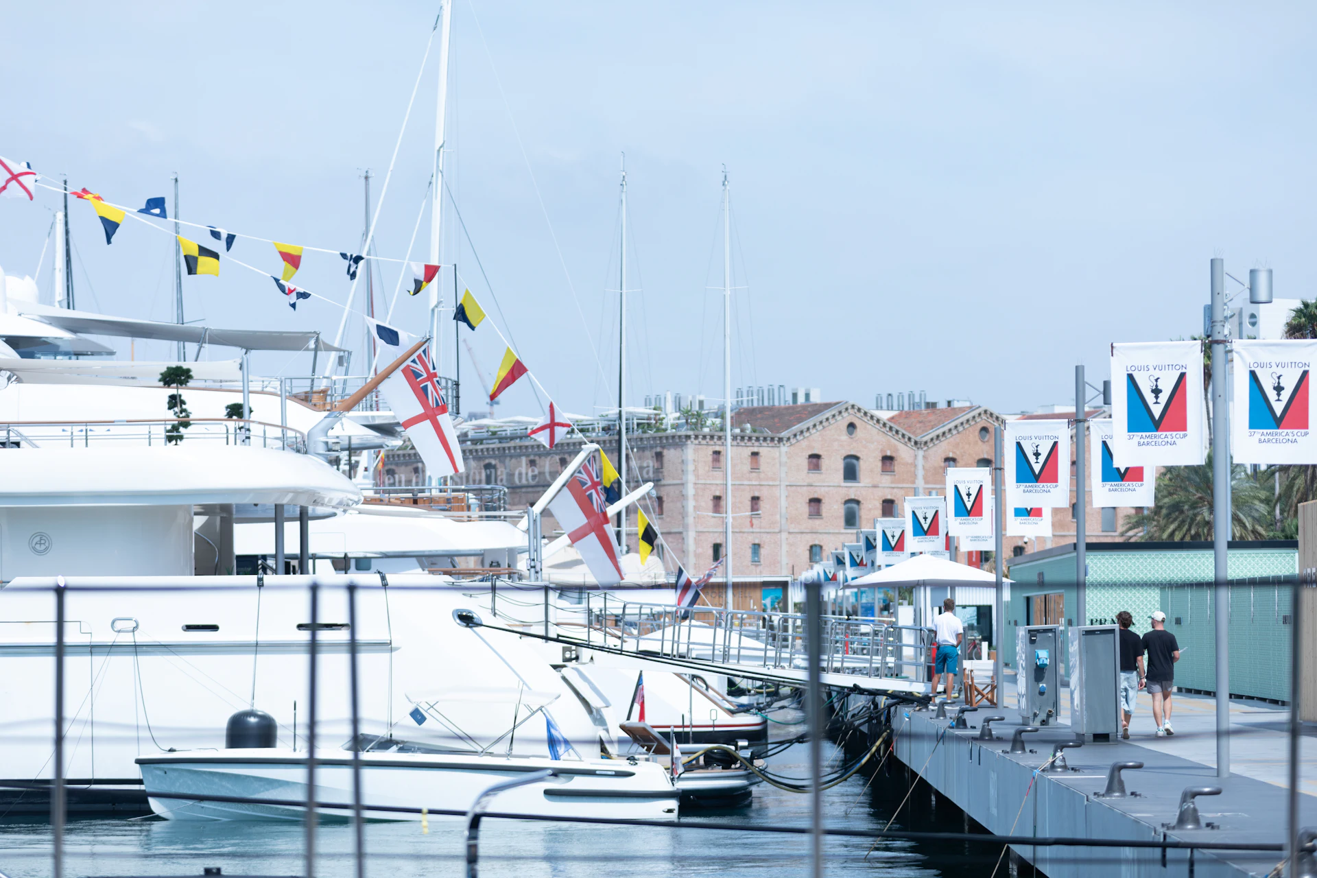 Superyates y veleros amarrados en Marina Port Vell con banderas náuticas y edificios históricos frente al mar en Barcelona.
