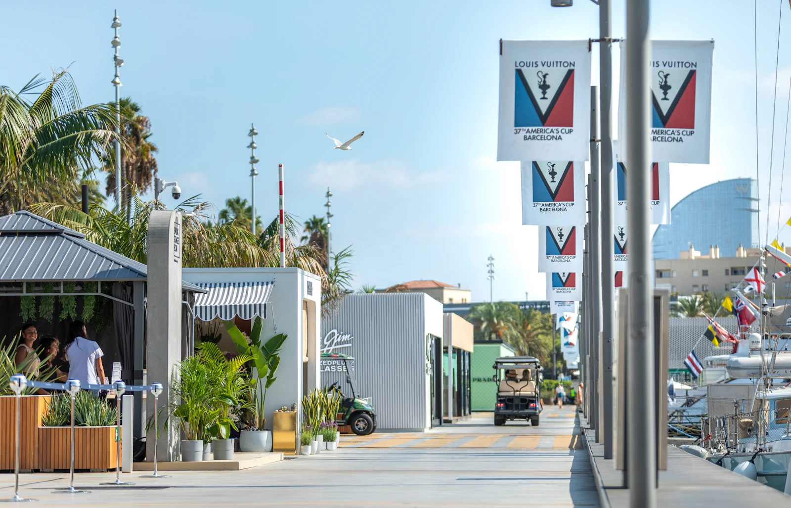 Marina promenade with America's Cup banners, palm trees, waterfront restaurants and moored boats on a sunny day.