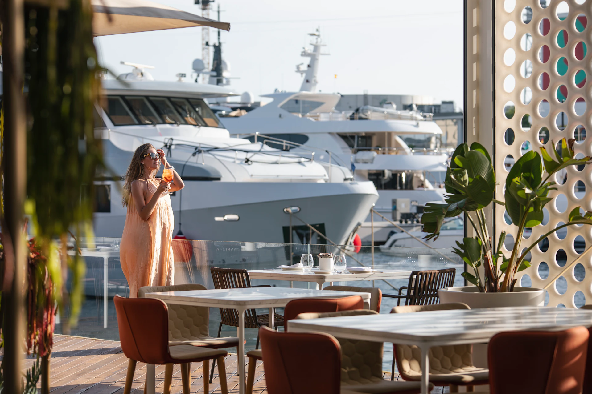 Woman in a light dress standing on a waterfront terrace overlooking docked superyachts.
