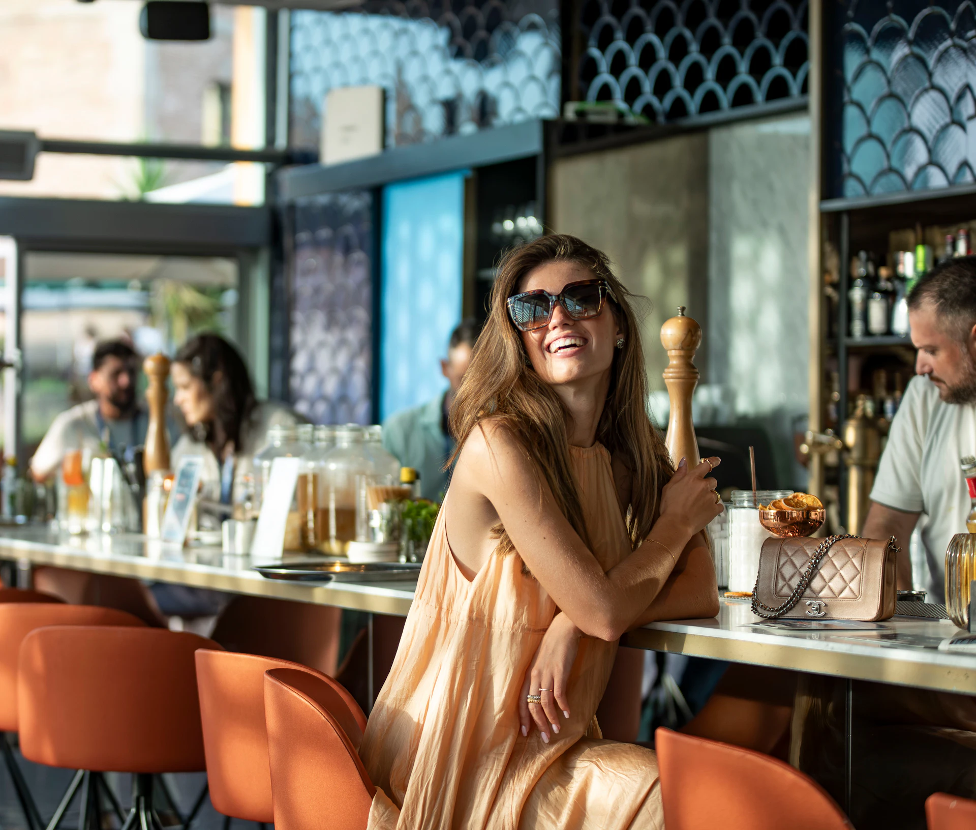 Mujer con gafas de sol y vestido beige sentada en la barra de un bar con un cóctel y un bolso.