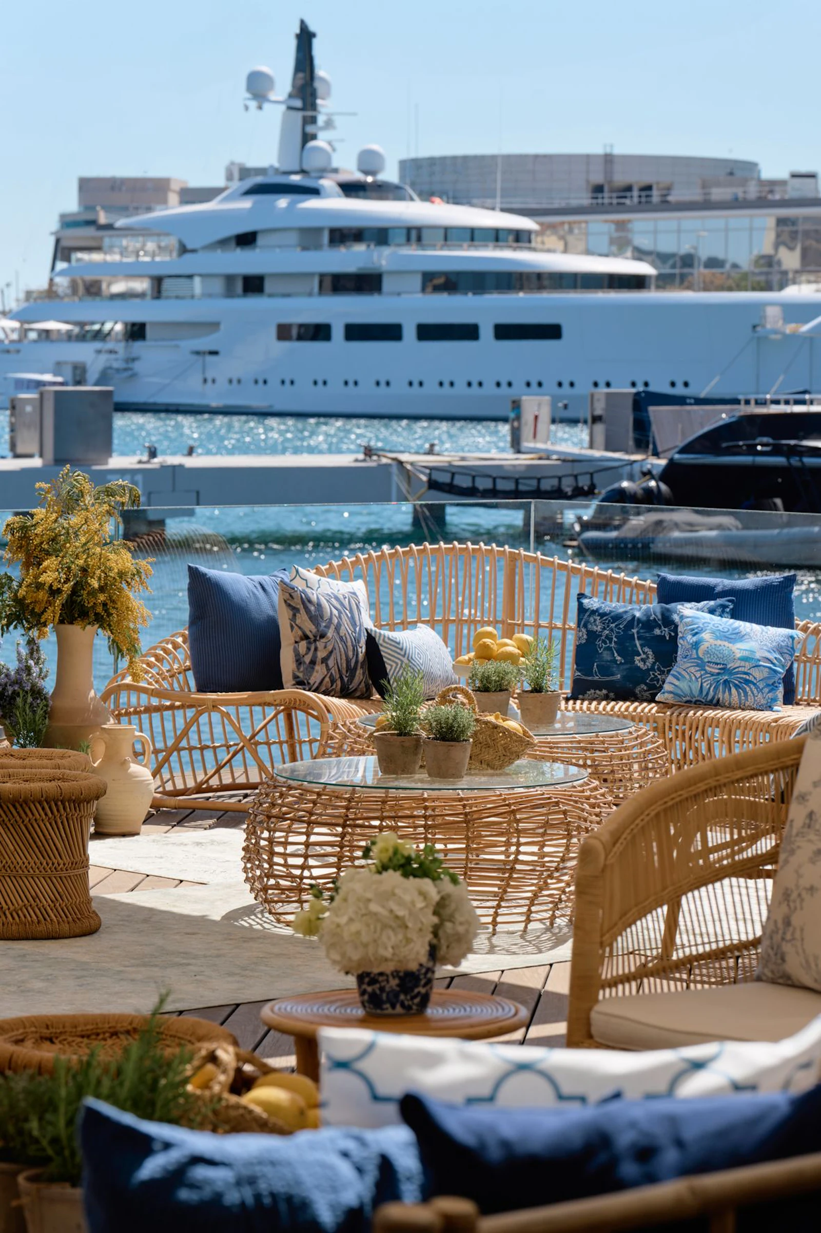 Woven seating area with cushions overlooking a large white superyacht moored at a marina.