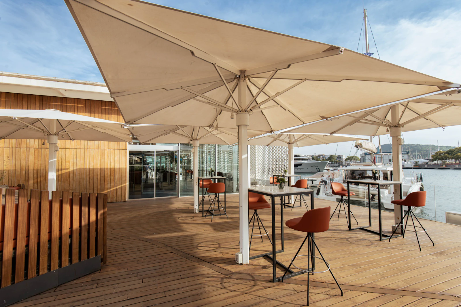 Waterfront terrace with large beige umbrellas, wooden decking, bar seating, and moored superyachts.
