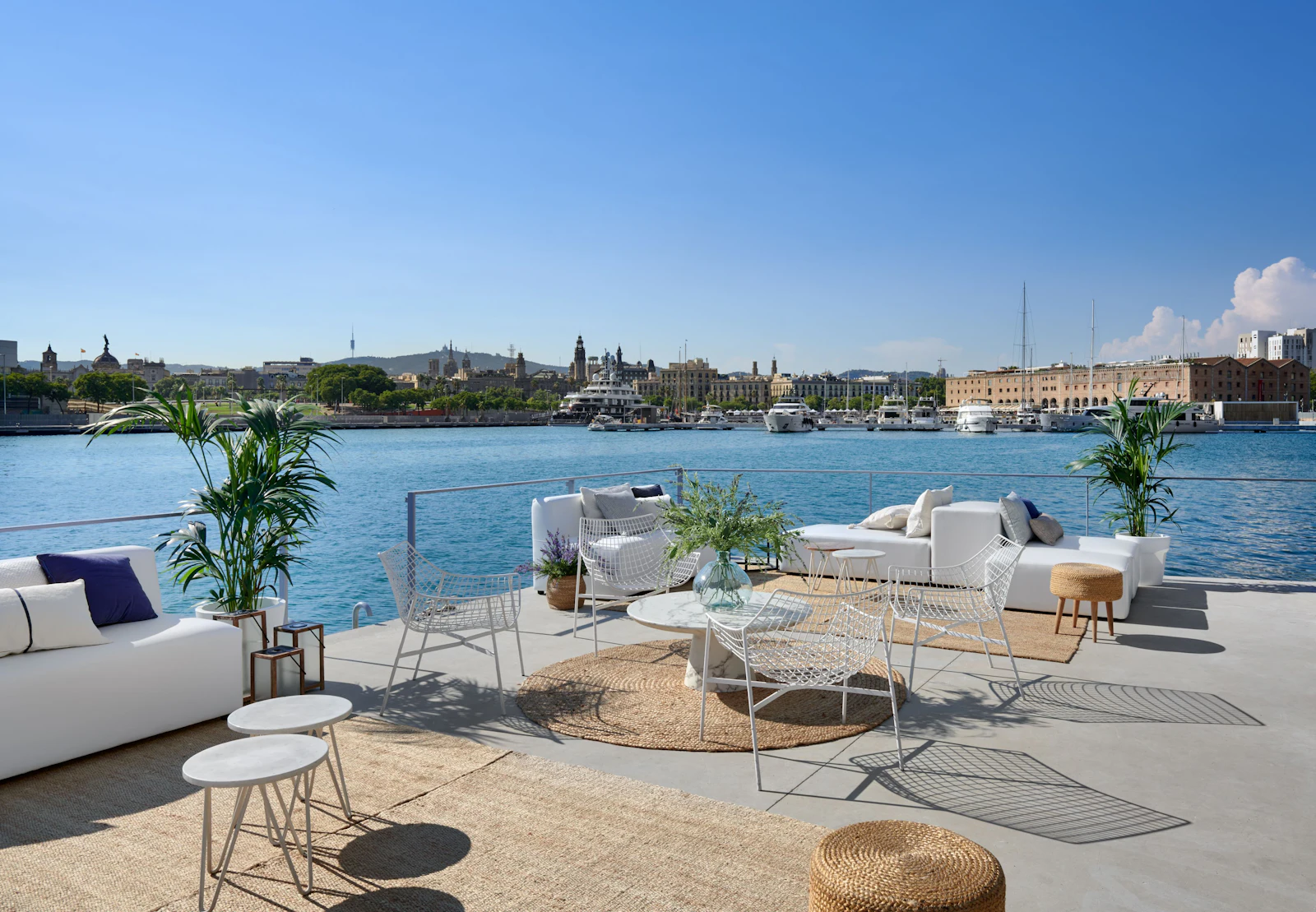 Waterfront lounge area with white furniture overlooking Barcelona's skyline and moored superyachts.