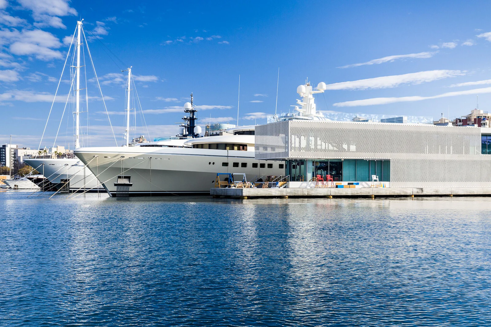 Gran superyate amarrado en el muelle de la marina junto a un edificio blanco moderno bajo un cielo azul despejado.