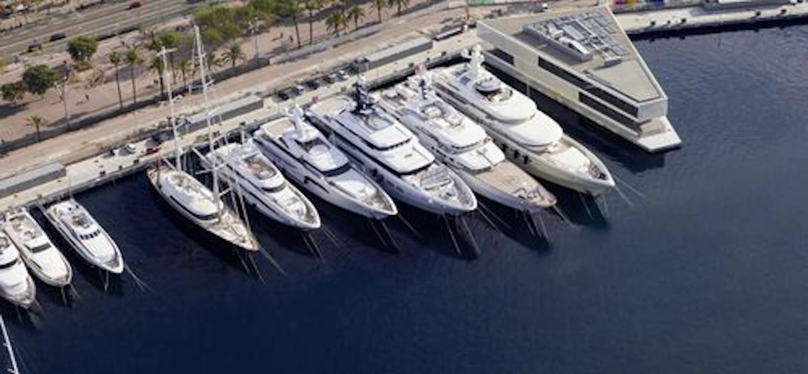 Multiple white and grey superyachts moored in a Barcelona marina with waterfront promenade and palm trees visible.