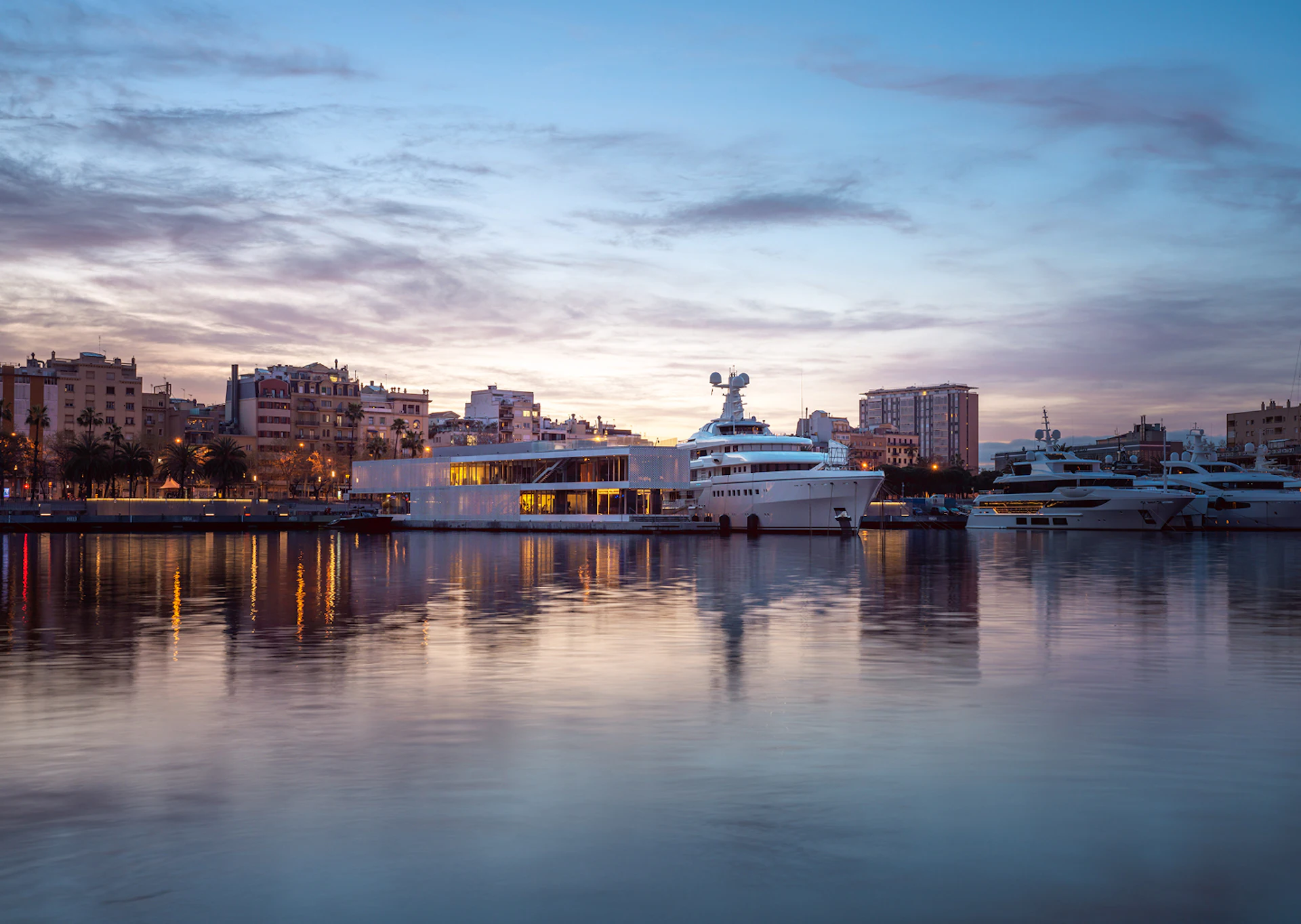 Superyachts moored at Barcelona marina at dusk with city skyline and illuminated waterfront buildings reflected in calm water.
