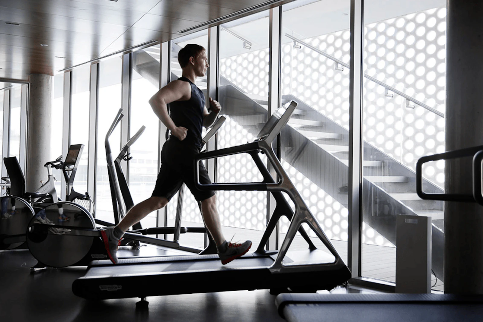 Man running on a treadmill in a gym beside large windows.