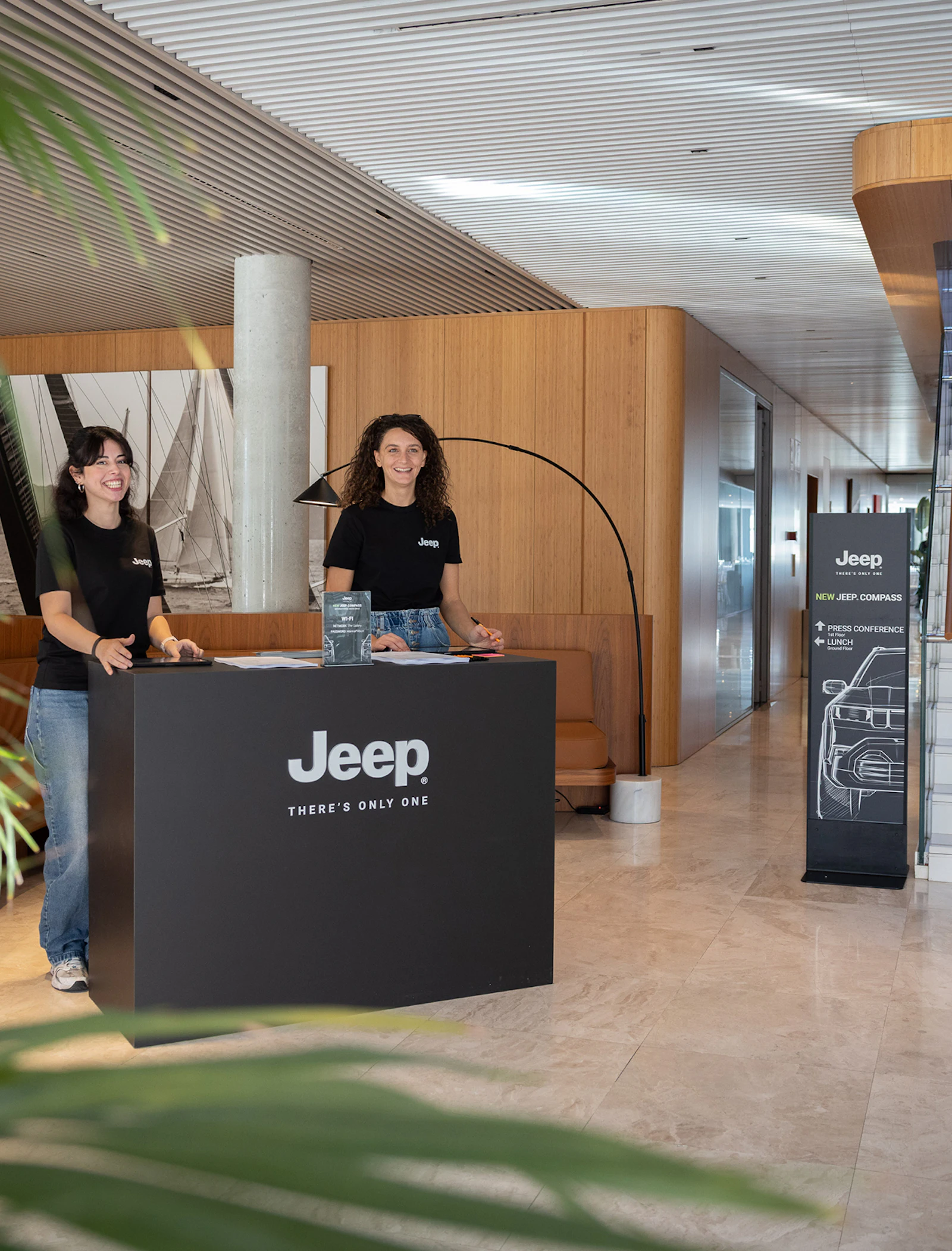 Two smiling women in Jeep shirts stand at a branded reception desk indoors.