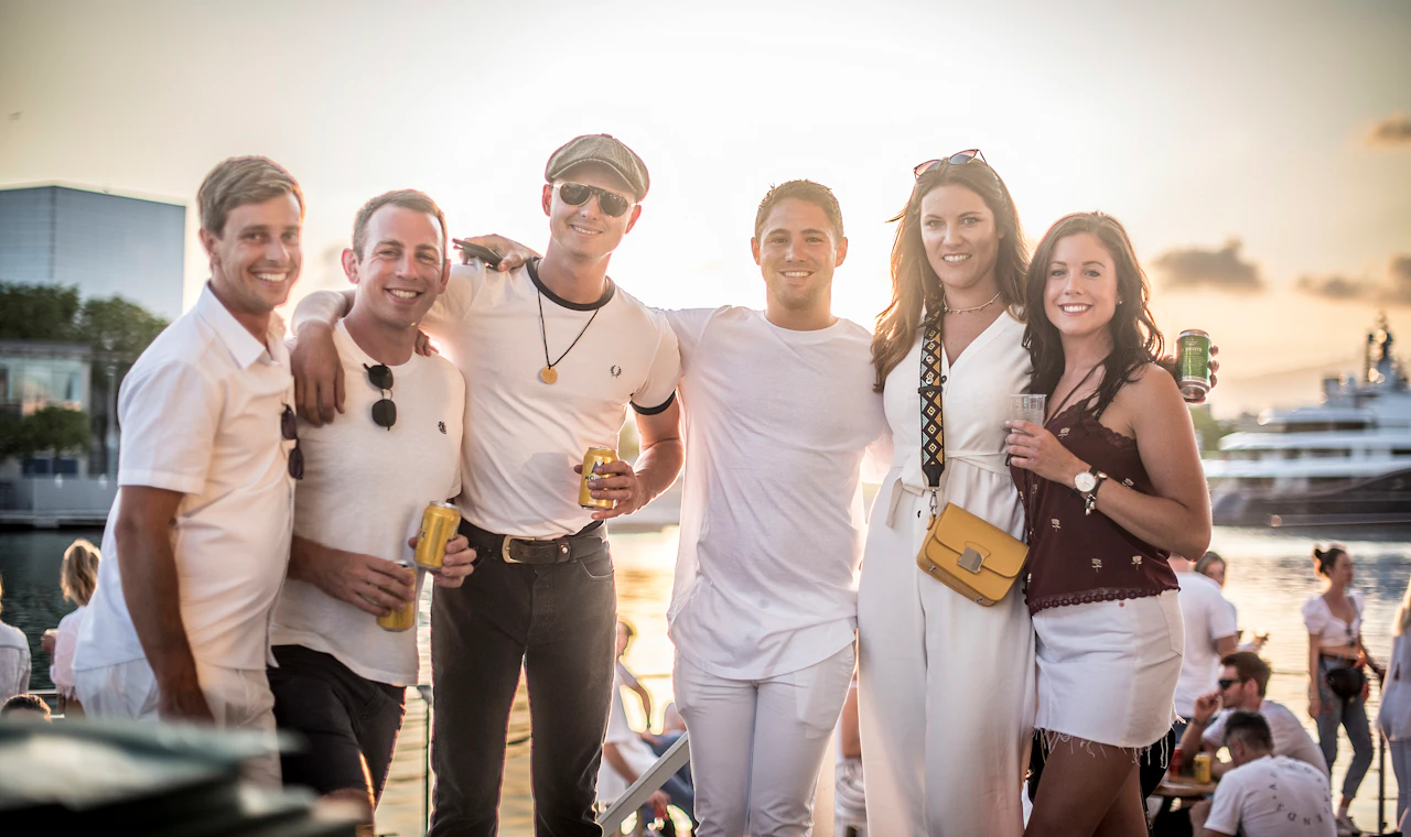 Group of friends with drinks posing at a waterfront event during sunset.