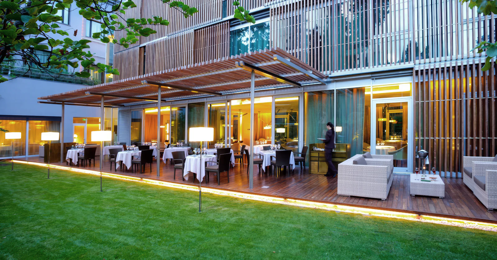 Outdoor terrace restaurant with set tables and lounge seating beside a lawn at dusk.