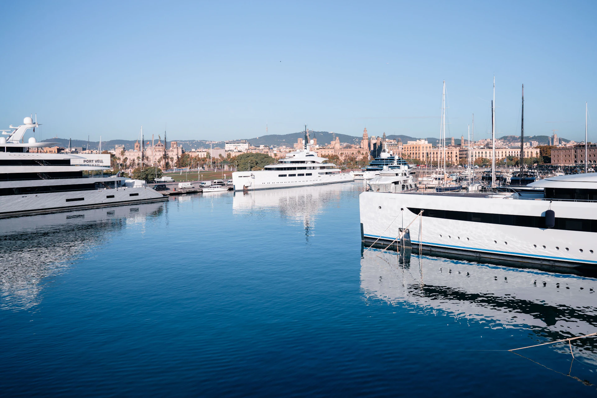 Superyachts moored at a marina with Barcelona's cityscape and mountains visible in the background.