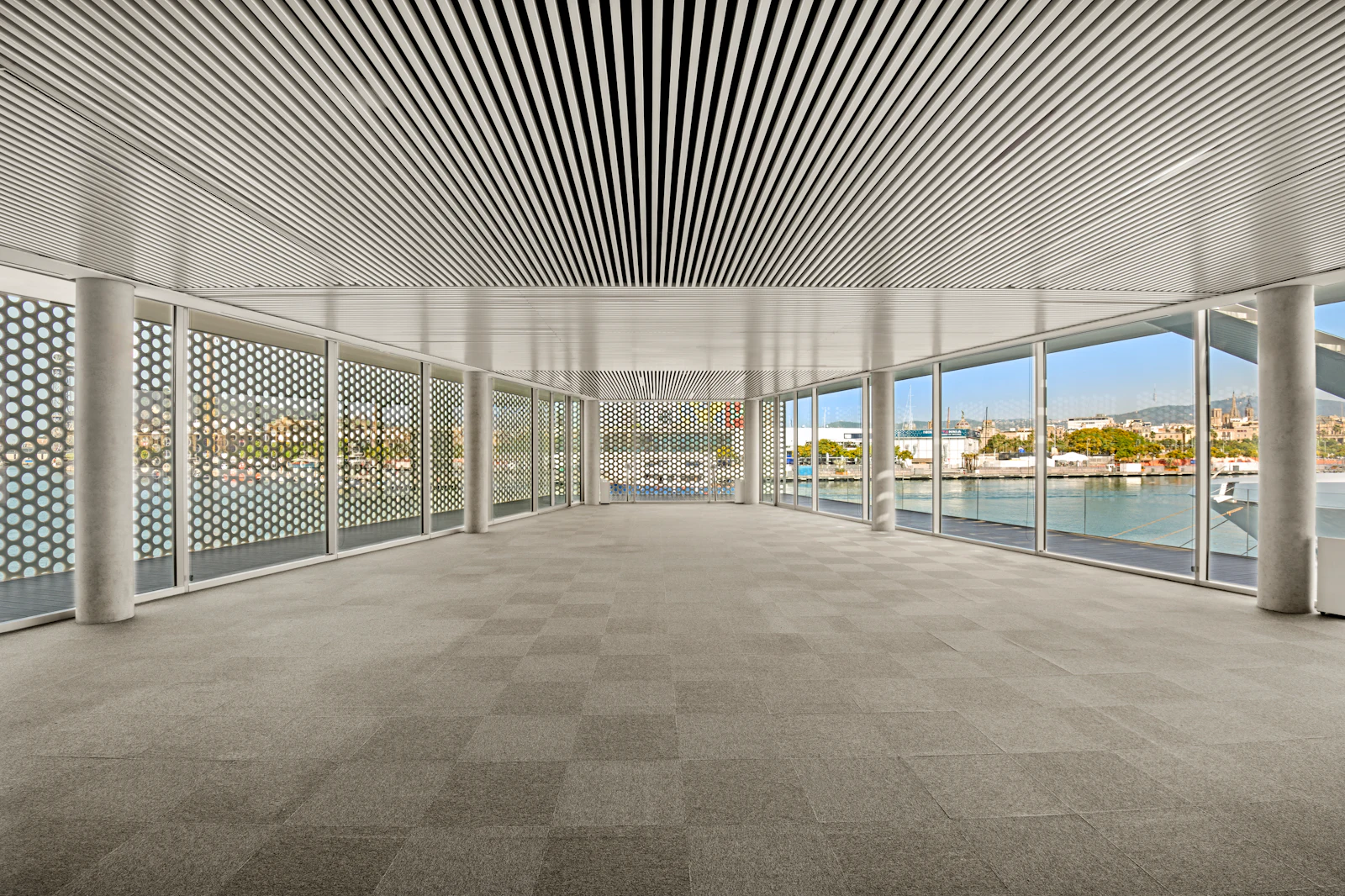 Espacio interior moderno y vacío con ventanales del suelo al techo con vistas a un puerto deportivo y al frente marítimo de Barcelona.