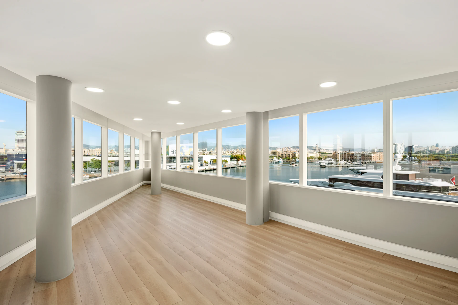 Empty modern interior space with large windows overlooking a marina and Barcelona cityscape.