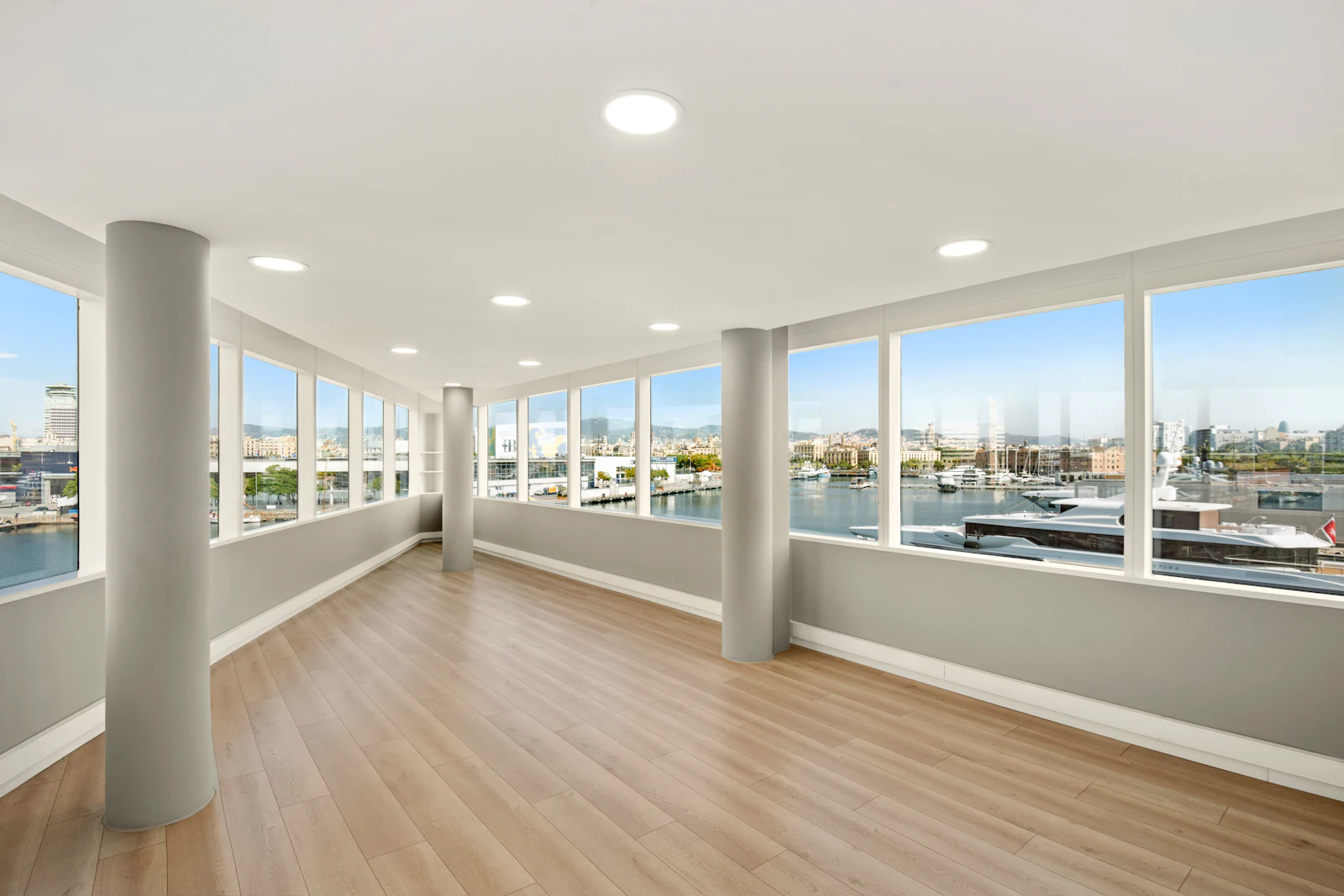 Empty modern interior space with wooden flooring, support columns, and expansive windows overlooking a marina and Barcelona cityscape.