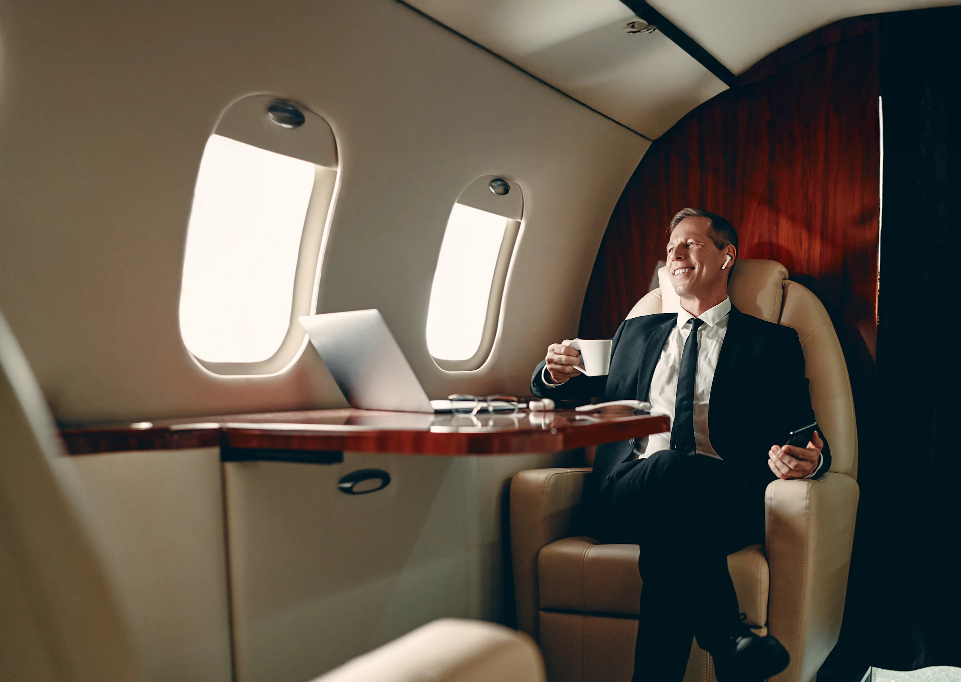 Smiling businessman in a suit holding coffee on a private jet.