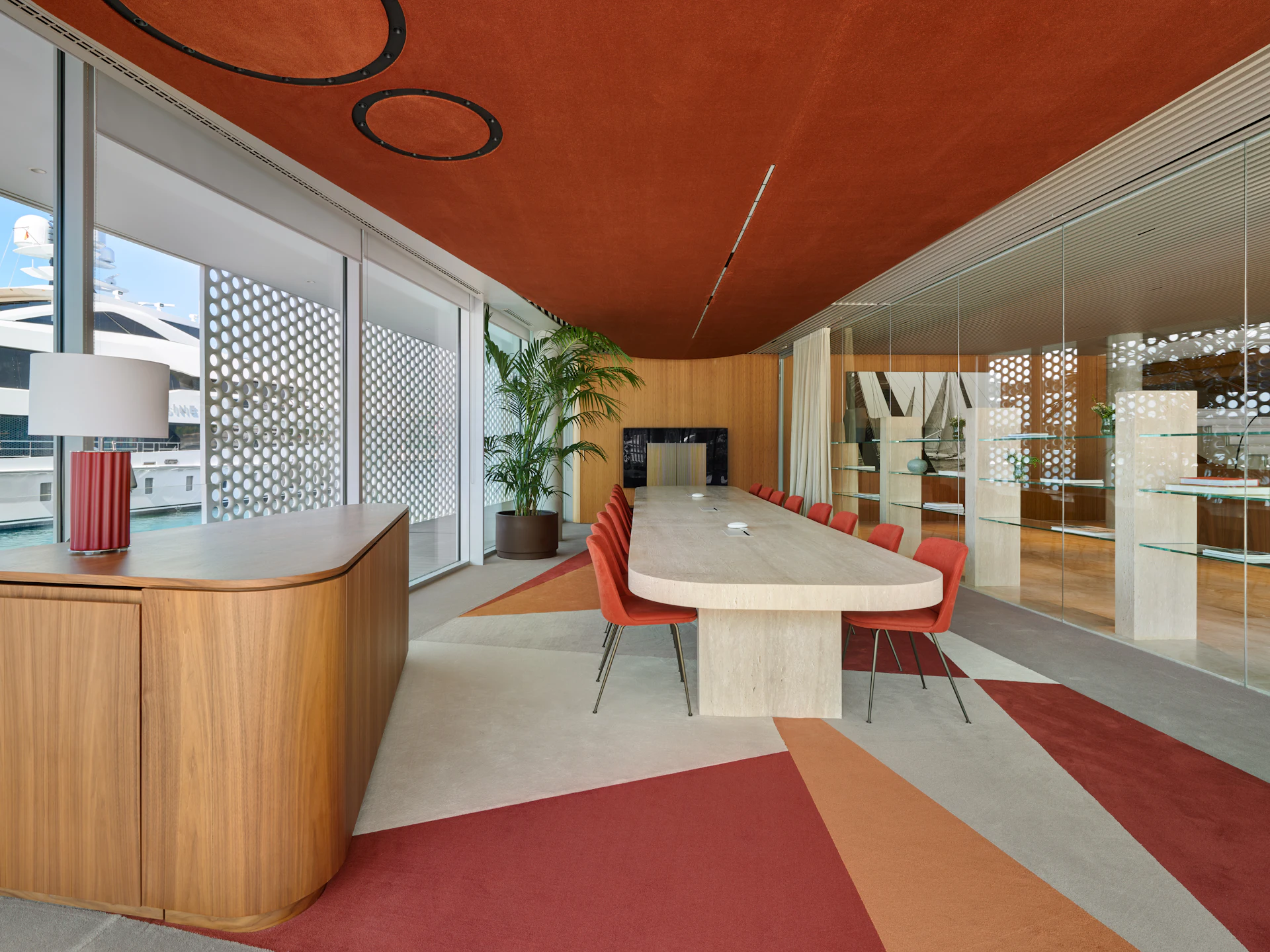 Modern marina lounge with red ceiling, wooden reception desk, conference table with red chairs, and harbor views through patterned glass doors.