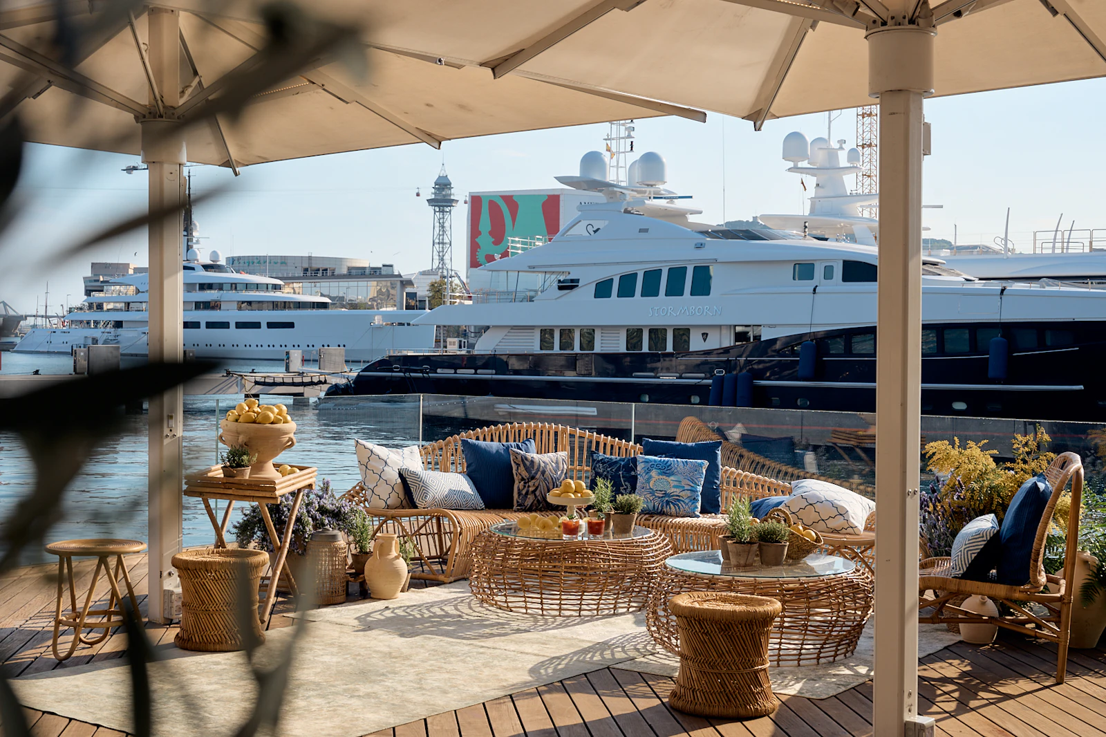 Waterfront lounge area with wicker furniture and umbrellas overlooking moored superyachts in Barcelona marina.