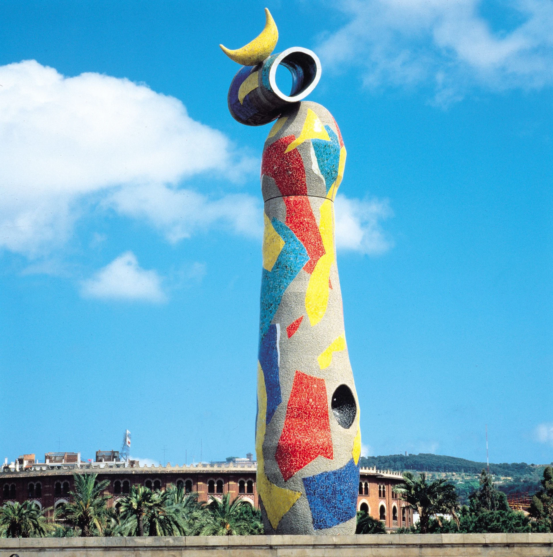 Colorful mosaic sculpture with moon and telescope top at Barcelona waterfront against blue sky.