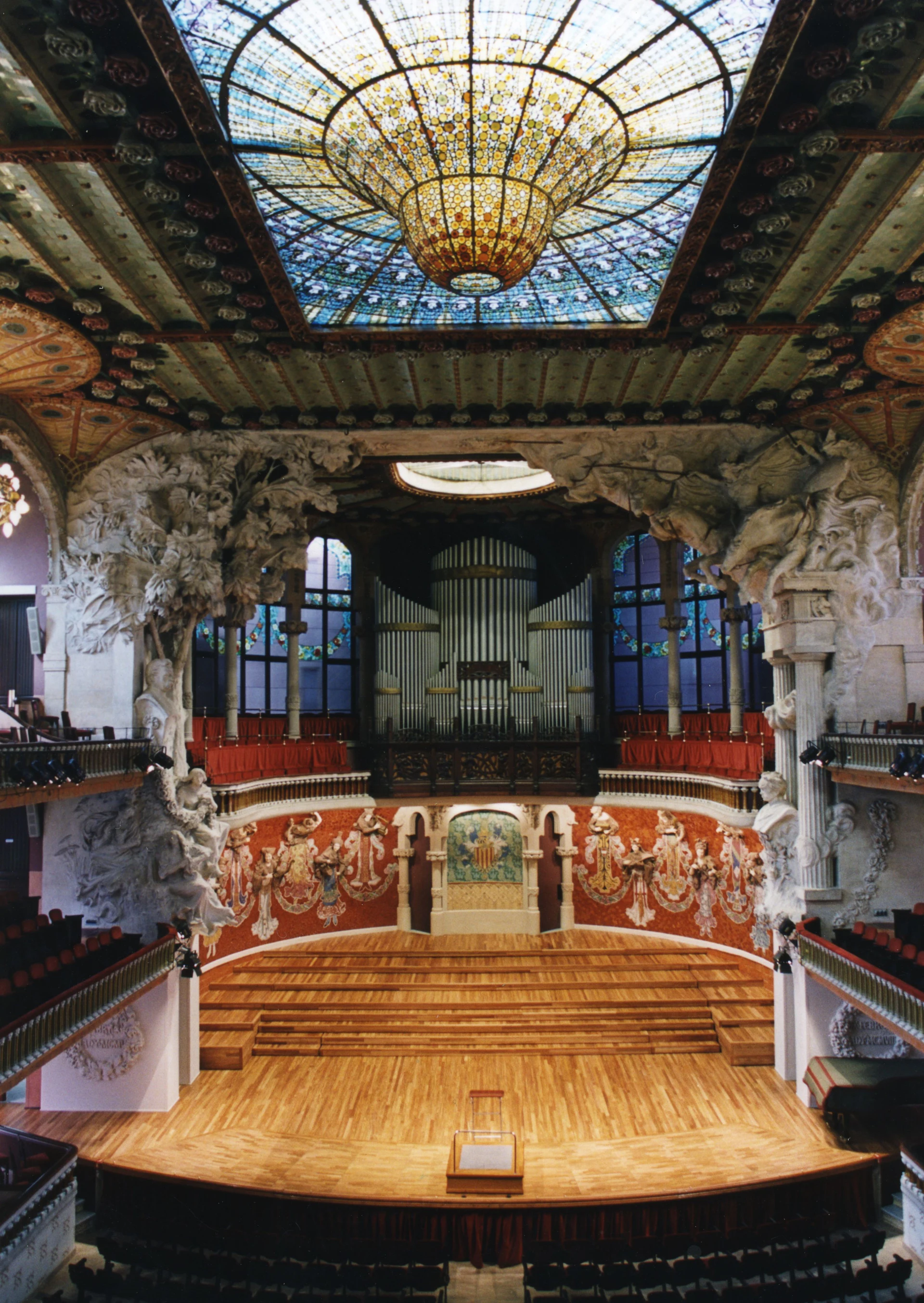 Ornate concert hall interior with stained glass ceiling, pipe organ, and decorative architectural details.