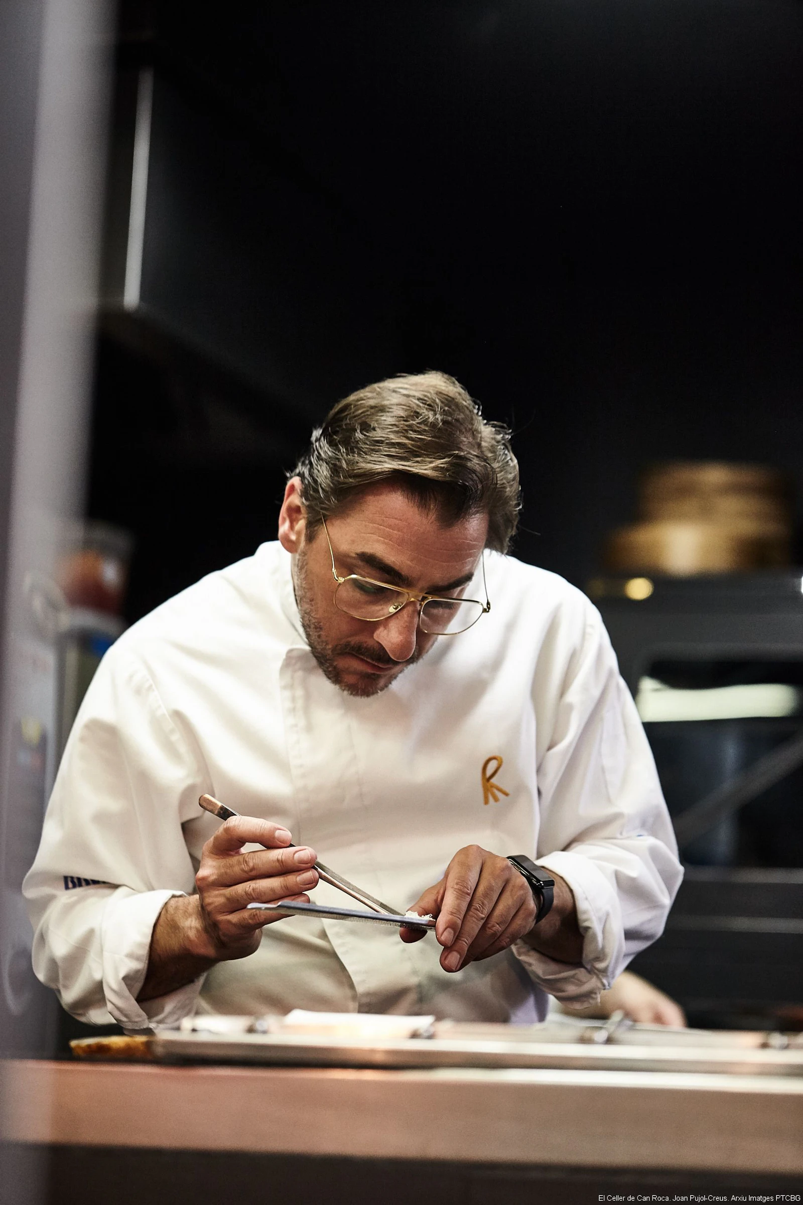 Chef in white chef's coat and glasses plating food with tweezers in a kitchen.