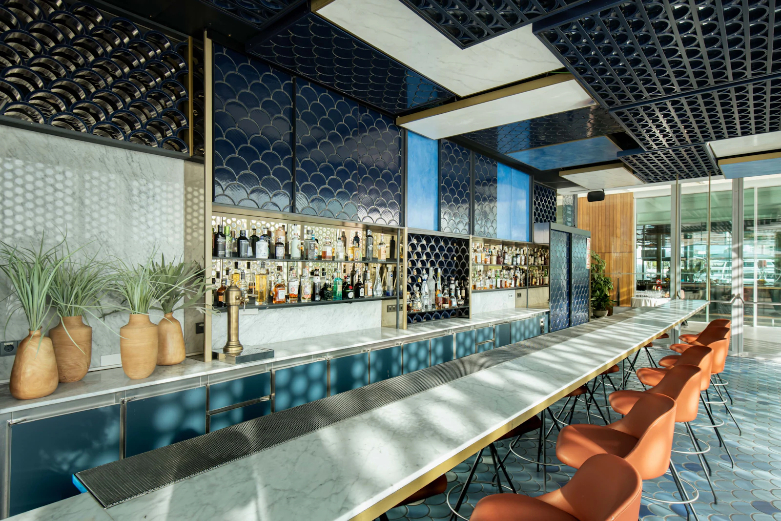 Modern bar with marble countertop, orange seating, decorative ceiling panels and potted plants.