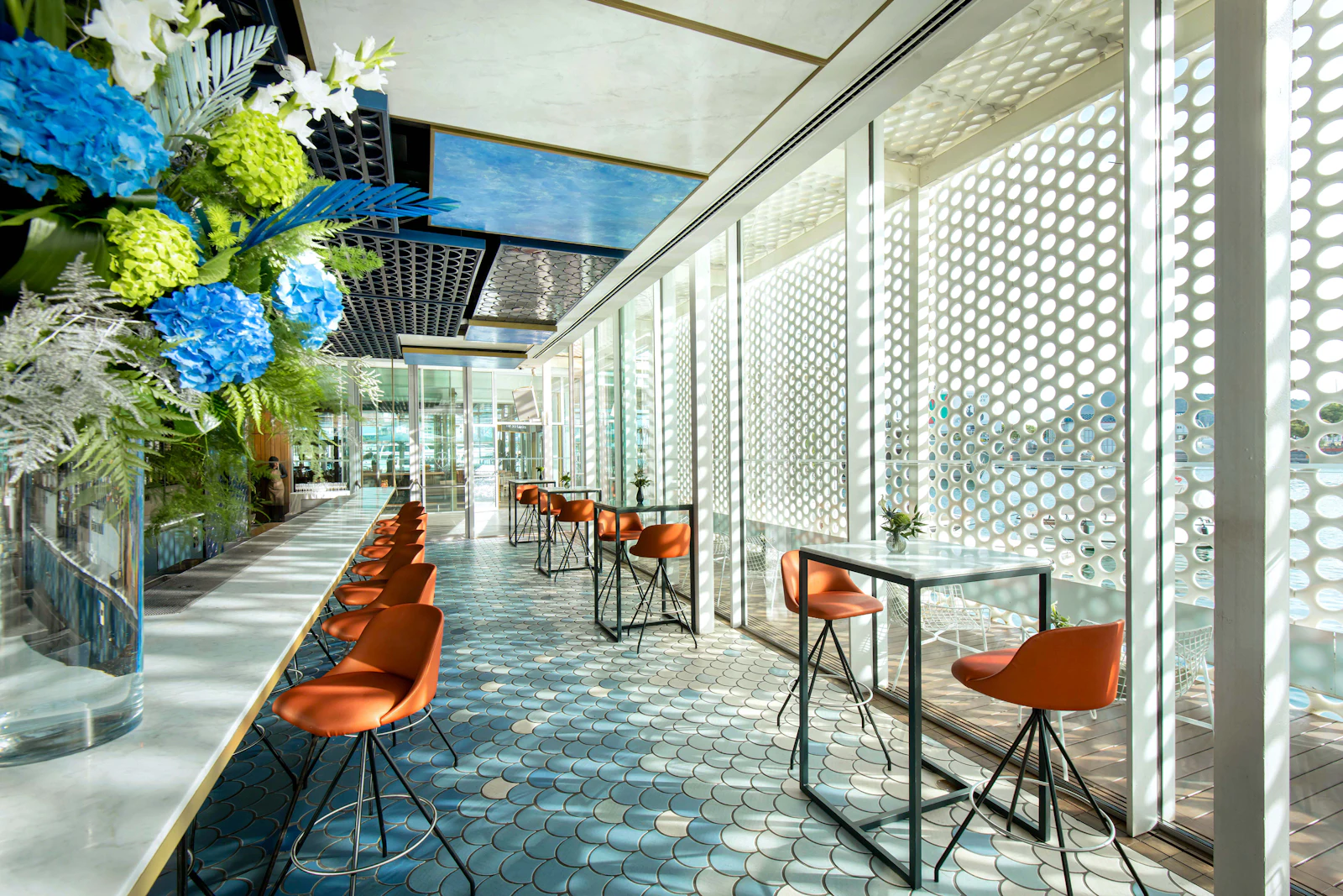 Modern café interior with orange stools, high tables and blue floral arrangement by perforated white screens.