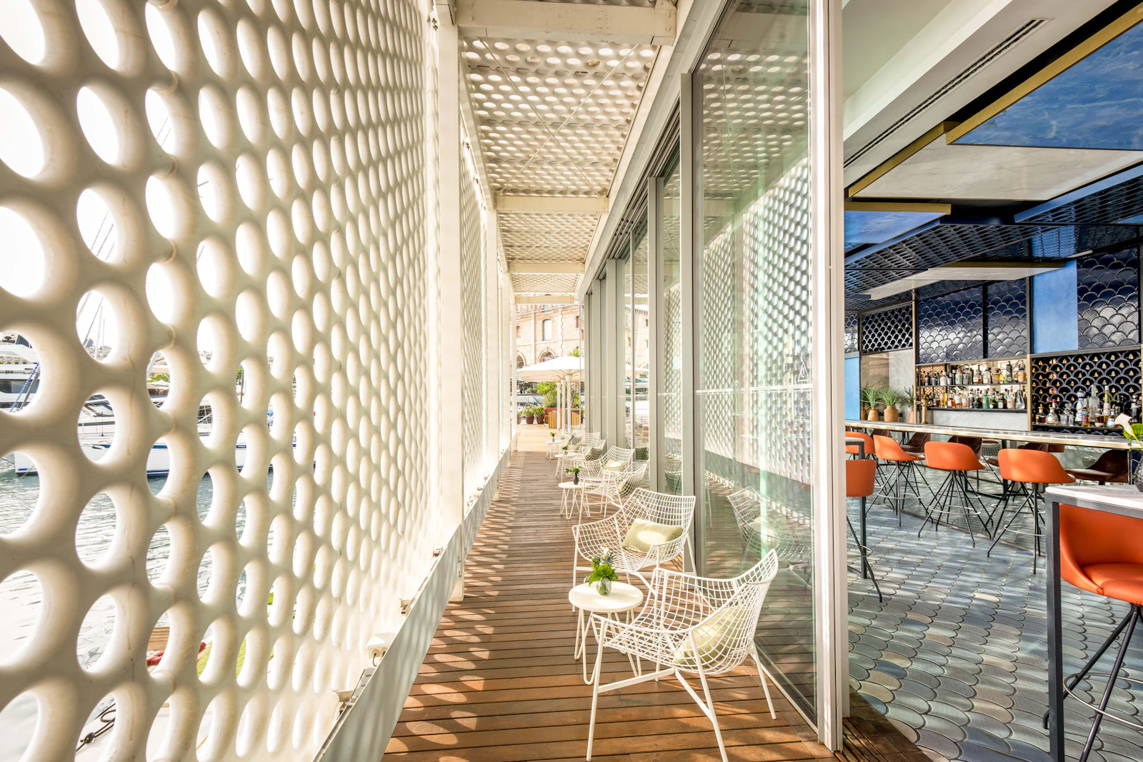Terrace with white wire chairs beside a perforated wall and glass-fronted bar interior.