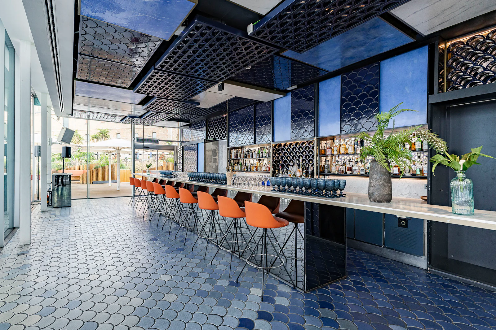 Modern bar with orange stools, blue tiled decor and stocked shelves.