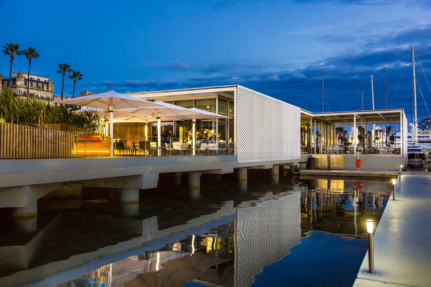 Waterfront restaurant with terrace umbrellas lit at dusk beside moored yachts and palm trees.