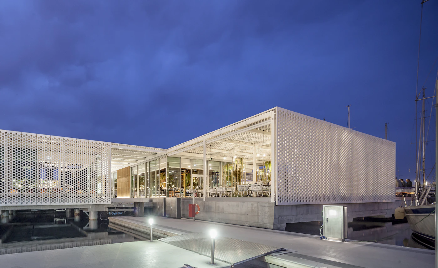 Illuminated glass restaurant with perforated white facade at the marina during twilight.