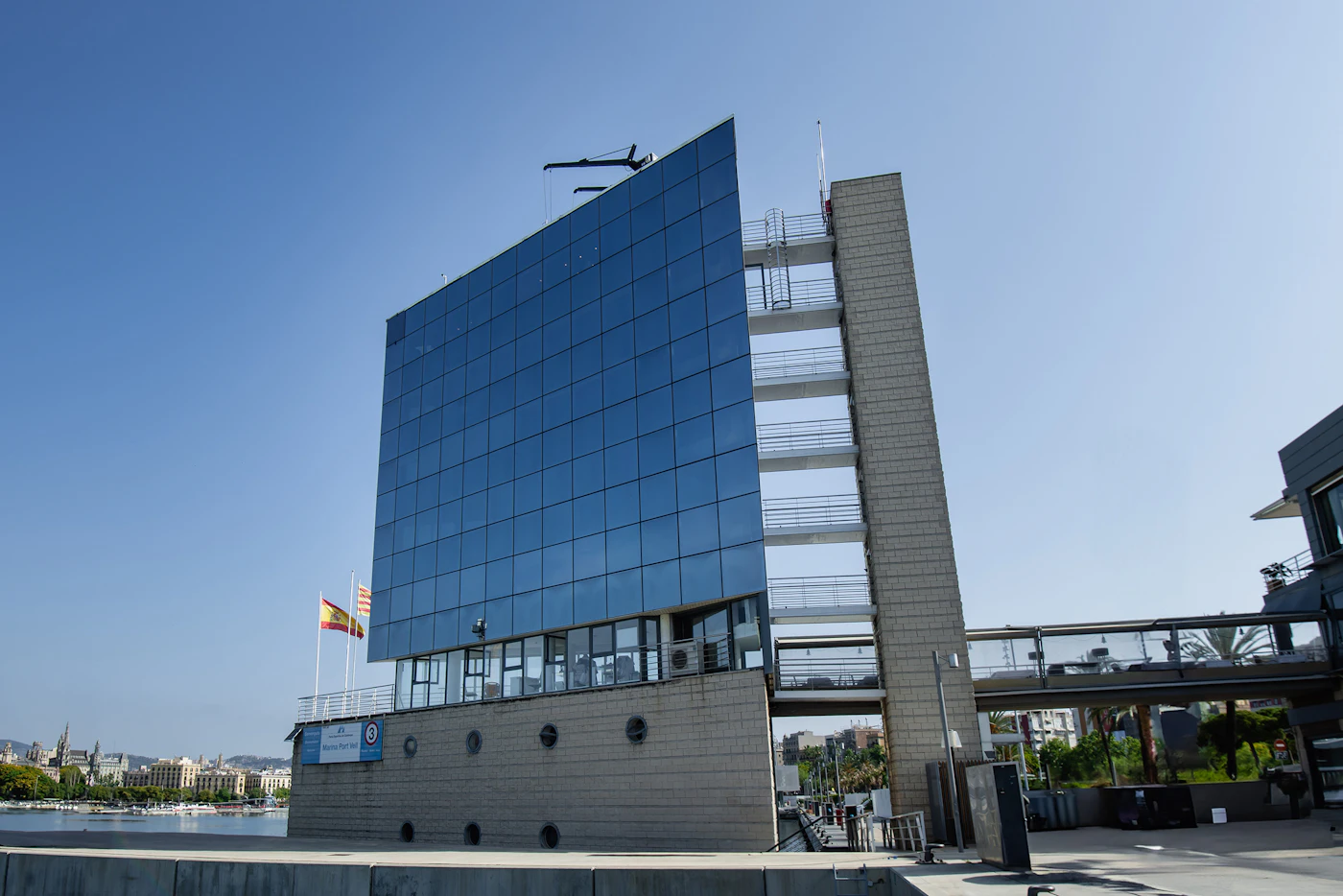 Modern glass-fronted marina building with Spanish and Catalan flags under a clear blue sky.