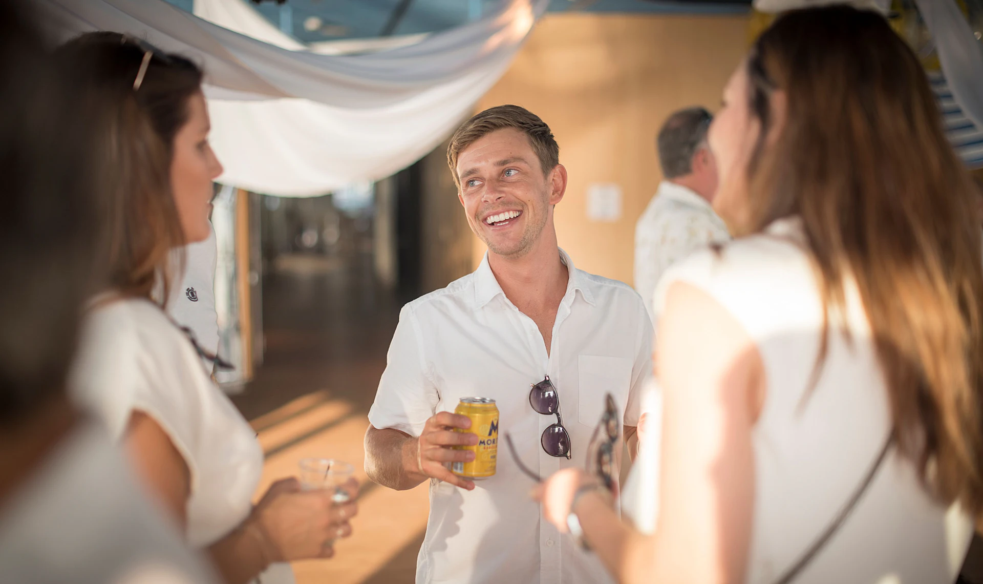 Hombre con camisa blanca sosteniendo una bebida conversa con colegas en un evento en interiores.