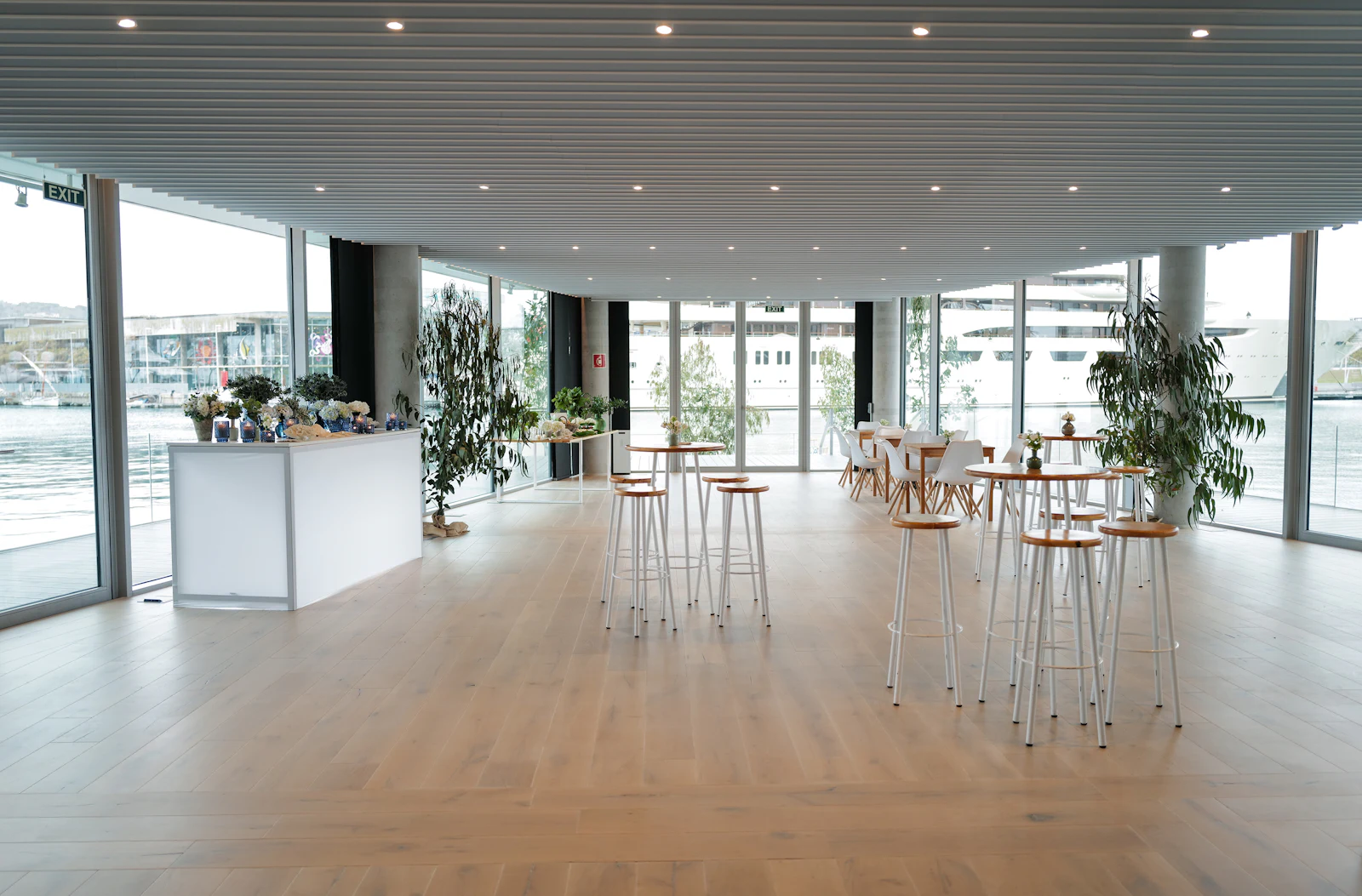 Espacio para eventos luminoso con mesas altas, taburetes y una barra con vistas al puerto deportivo.