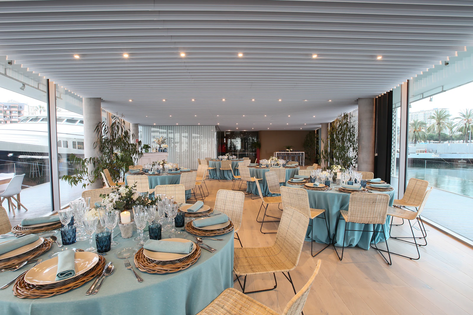 Espacio para eventos dispuesto con mesas redondas con mantelería azul y sillas de ratán junto a las ventanas del puerto deportivo.
