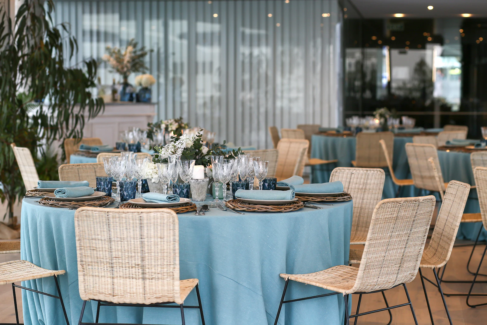Mesas redondas con mantelería azul claro, sillas de mimbre y centros florales dispuestas para un evento.