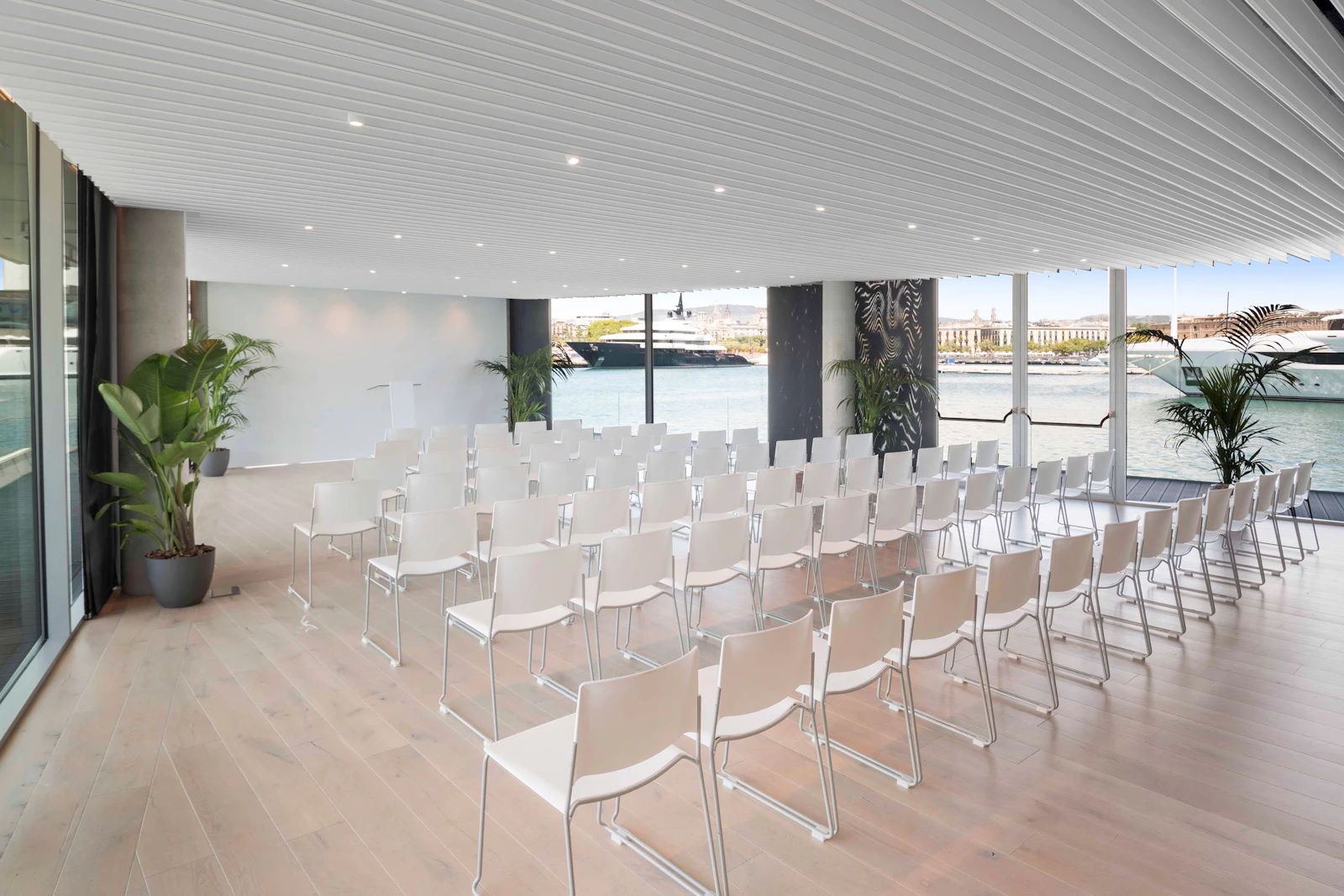 Filas de sillas blancas en una sala de eventos con vistas al puerto y yates.