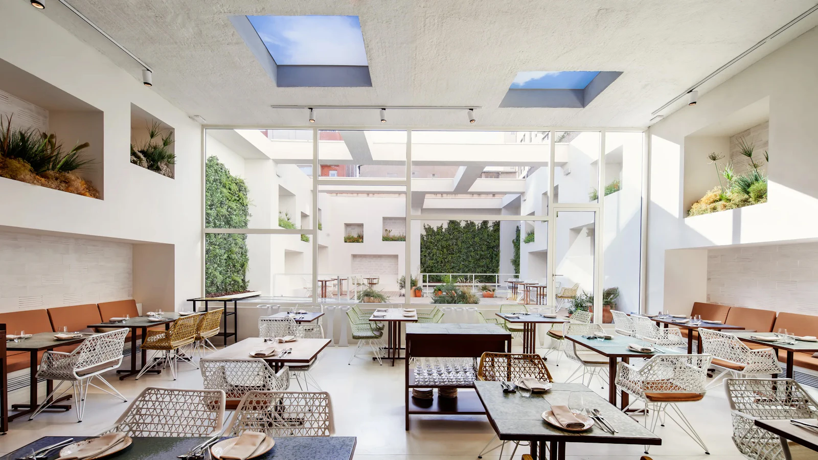 Bright restaurant interior with woven chairs, set tables, skylights and large windows overlooking greenery.