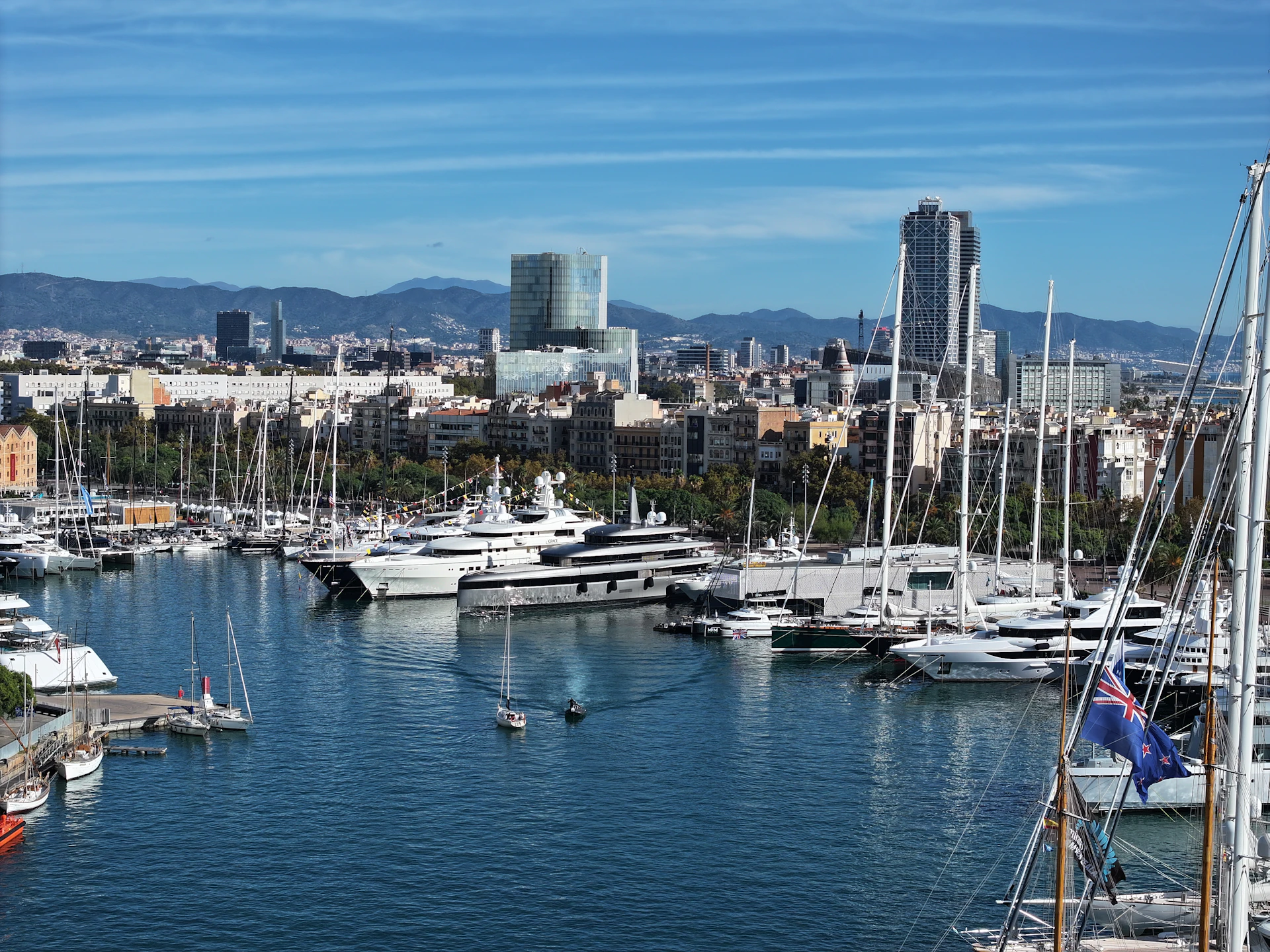 Superyachts and sailboats moored in Barcelona's waterfront marina with city skyline and mountains in background.