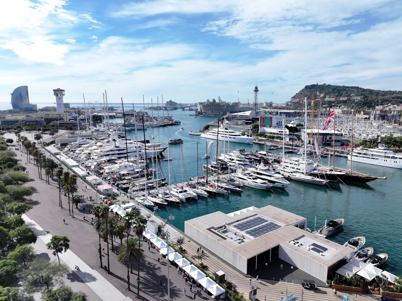 Marina filled with moored superyachts and sailboats, Barcelona waterfront with palm trees and city skyline visible.