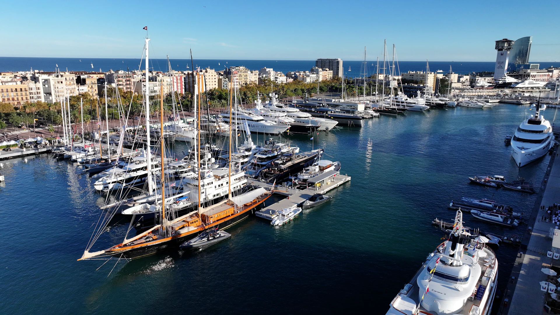 Superyates y veleros amarrados en la marina de Barcelona con el mar Mediterráneo y el perfil urbano de la ciudad al fondo.