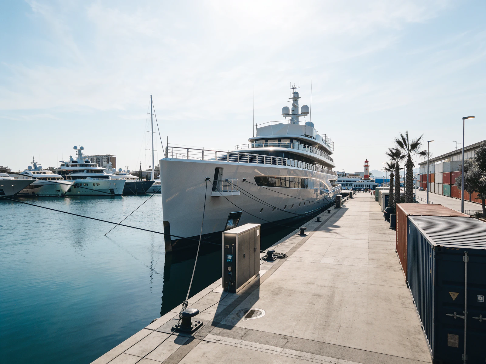 Large superyacht moored along a marina dock with palm trees and a lighthouse nearby.