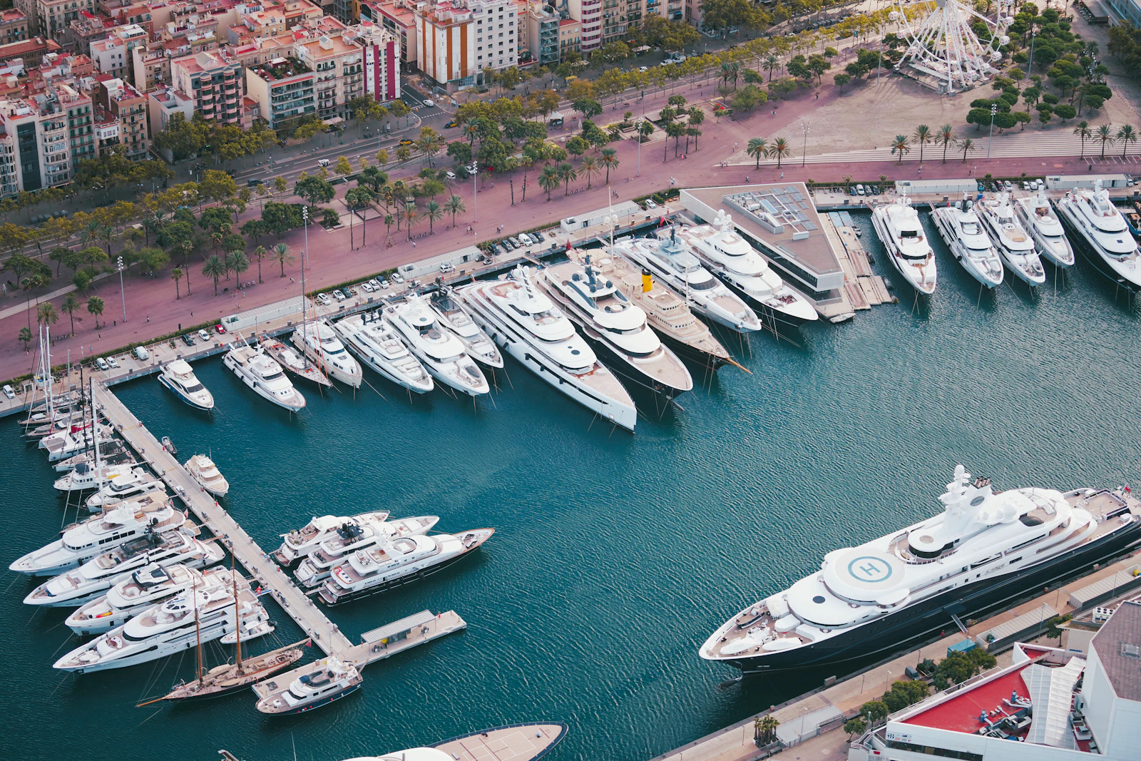 Aerial view of superyachts docked at a marina beside a palm-lined promenade and city buildings.