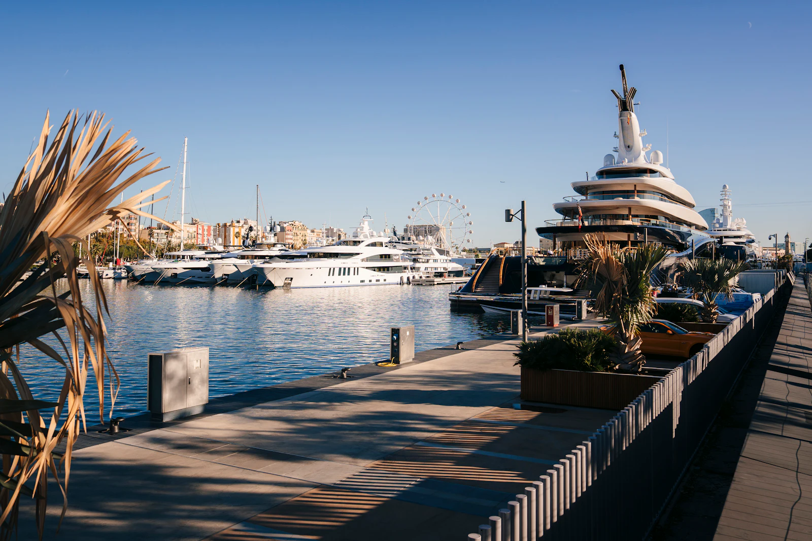 Superyachts moored along a marina promenade with a Ferris wheel in the background.