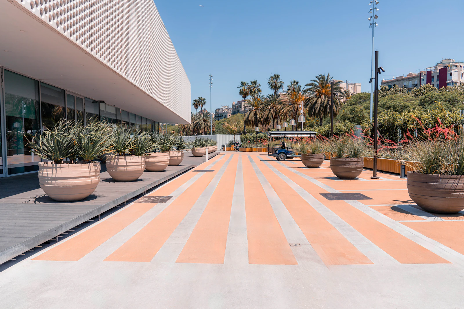 Striped orange and grey plaza lined with large planters beside a modern building and palm trees.