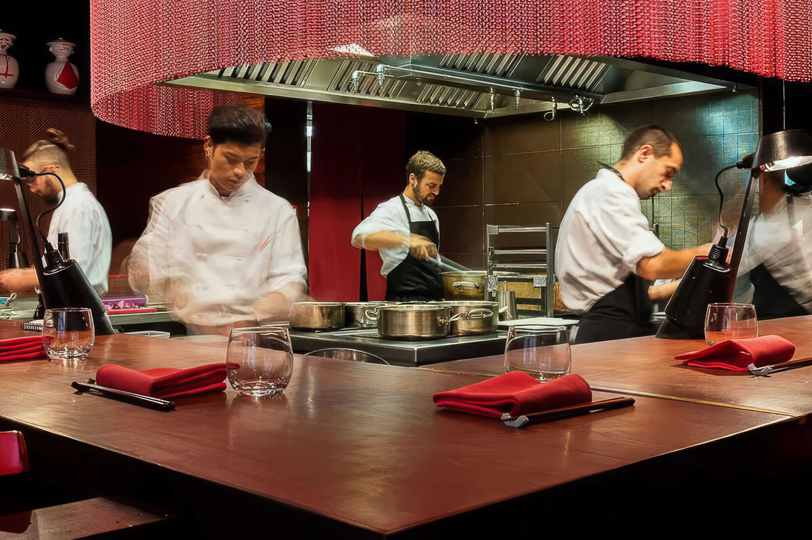 Chefs cooking in an open kitchen behind a counter set with glasses and red napkins.