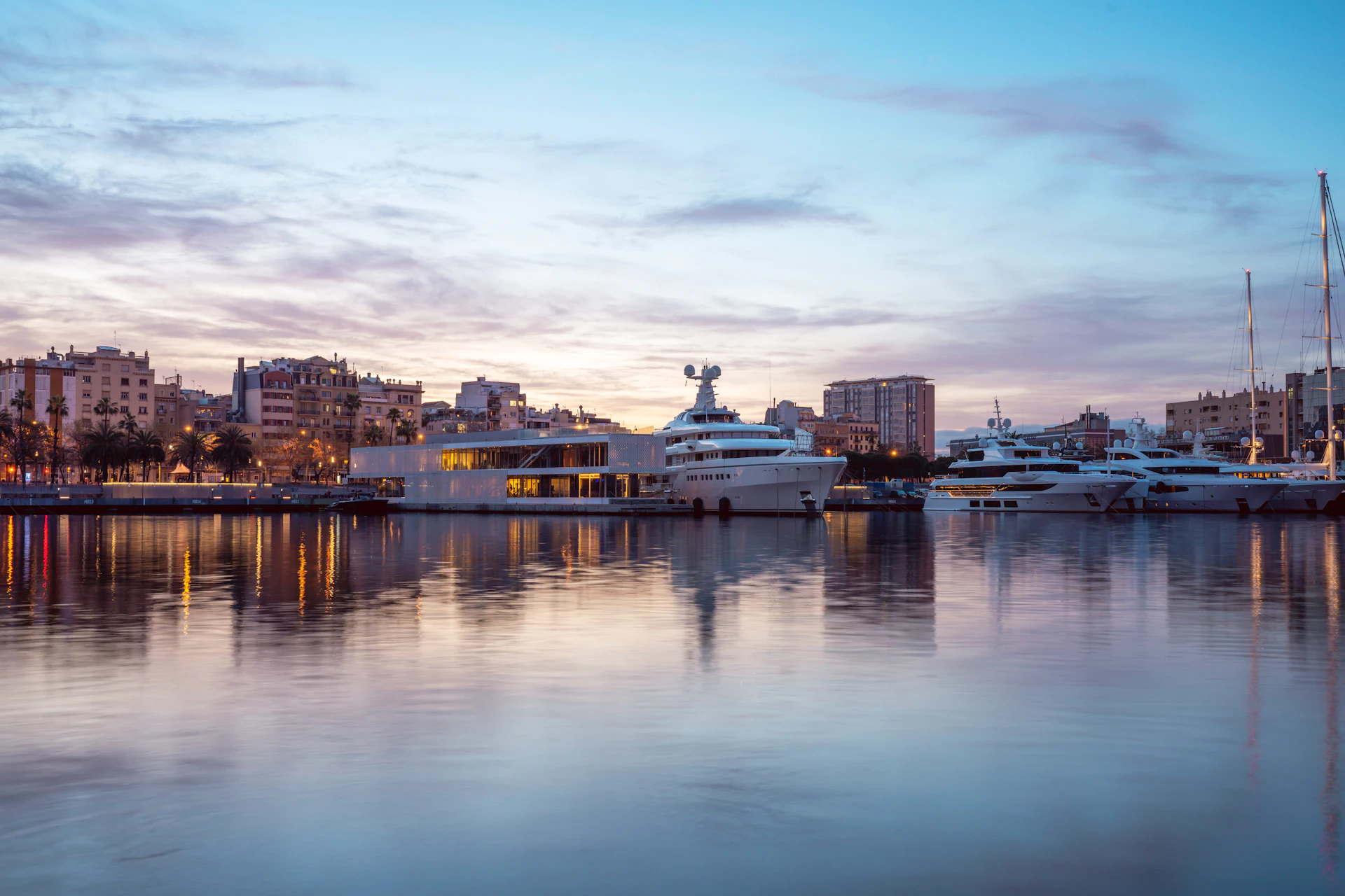 Superyachts moored at a marina at dusk with Barcelona waterfront buildings reflected in calm water.