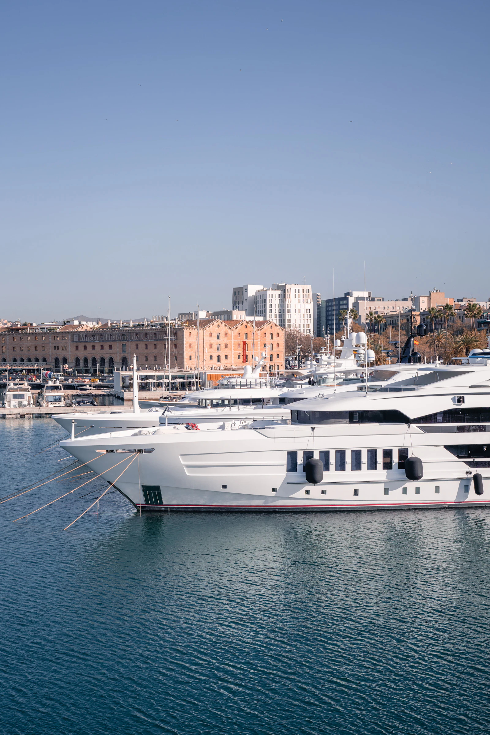 White superyacht moored at Barcelona's Port Vell marina with historic waterfront buildings in background.