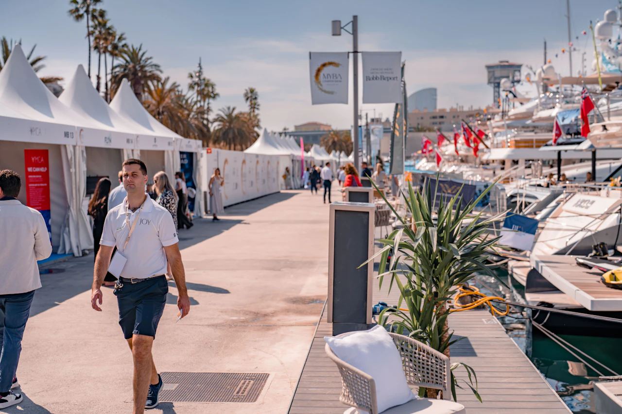 Man walking through marina promenade with white event tents, moored yachts, and palm trees under clear sky.